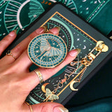 A close-up of a hand holding a round celestial-themed magnetic kindle grip with a luna moth charm attached by a chain. The design features teal, gold, and cream moon and star motifs, with a matching patterned surface visible in the background. By The Quirky Cup Collective