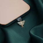 A Luna Moth dust plug design by The Quirky Cup Collective is plugged into a phone on a deep green fabric background. The gold Night luna moth design (as part of the romantasy kindle decor release) featured green detailing.