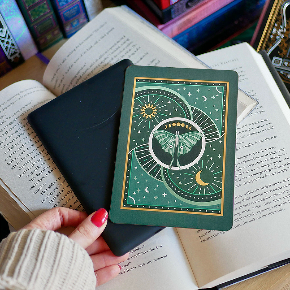 Image shows a hand gently touching a kindle and a kindle sticker insert sitting on top of the device. They sit on a bed of open books. The teal, gold and white design shows a luna moth surrounded by moons, stars and decorative embellishments. 