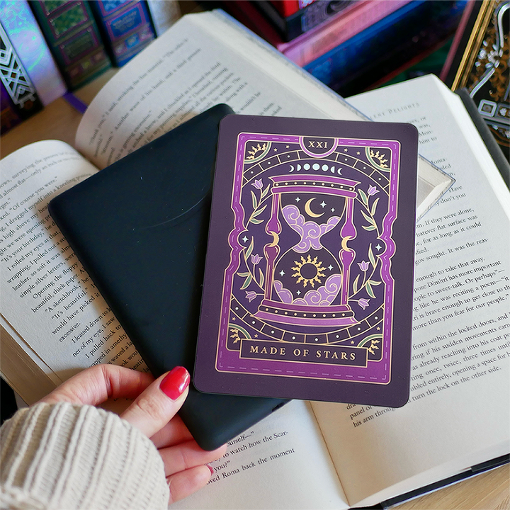 Image shows a hand gently touching a kindle and a kindle sticker insert sitting on top of the device. They sit on a bed of open books. The tarot inspired purple designs shows an hourglass with clouds, stars sun and a moon surrounded by a floral wreath surrounded by moons, stars and celestial decorative embellishments. 