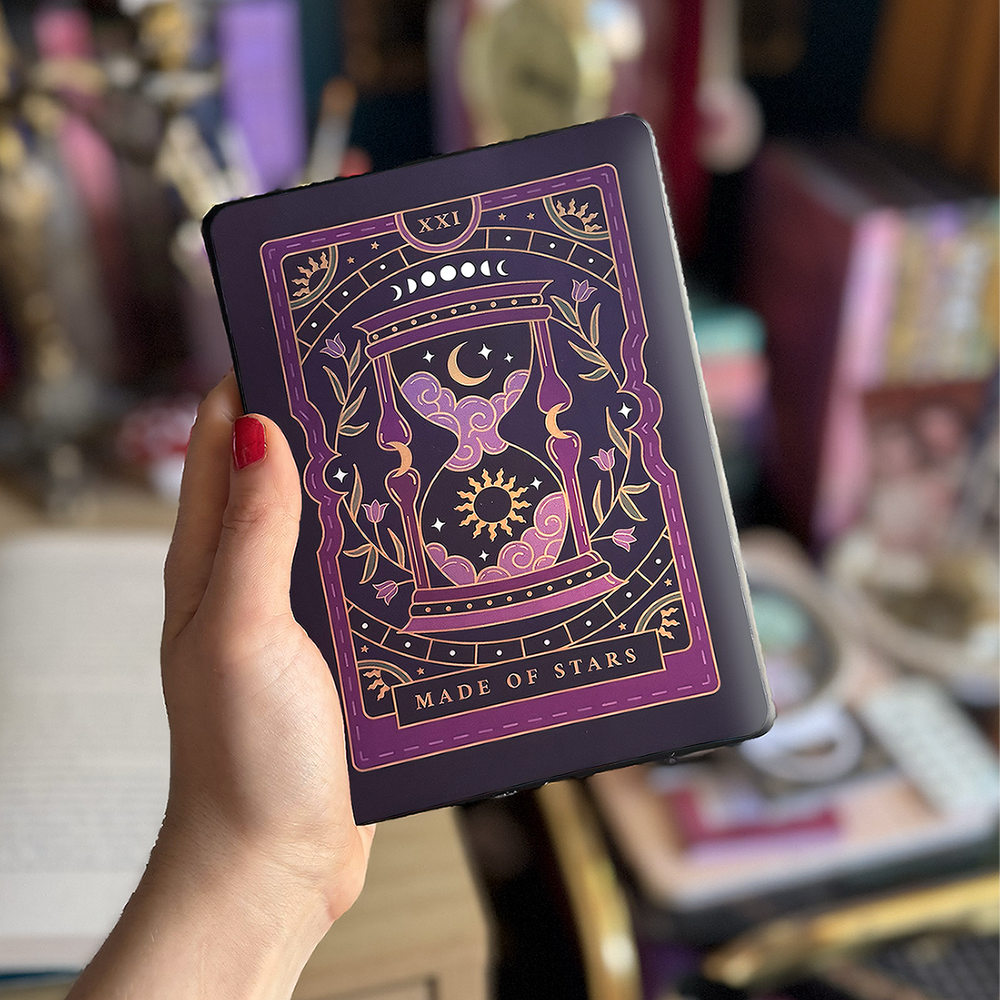 Image shows a hand holding a kindle showing off a kindle sticker insert that is placed on the back of the device. The tarot inspired purple designs shows an hourglass with clouds, stars sun and a moon surrounded by a floral wreath surrounded by moons, stars and celestial decorative embellishments. 