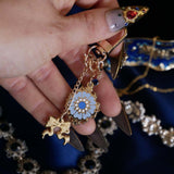 A hand holds The Quirky Cup Collective Moncherie charmlet, an accesory with  a series of charms attached to a central clip. The design is gold and features a bow charm, a blue gem and a enamel element of a floral motif in a decorative french inspired style in shades of blue, cream and gold. The french inspired eReader accessory is held by a hand with intricate gold nails in front of a blue fabric background.