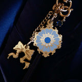 A detail shot of The Quirky Cup Collective Moncherie charmlet, an accessory with  a series of charms attached to a central clip. The design is gold and features a bow charm, a blue gem and a enamel element of a floral motif in a decorative french inspired style in shades of blue, cream and gold. The french inspired eReader accessory is placed on a royal blue fabric background.