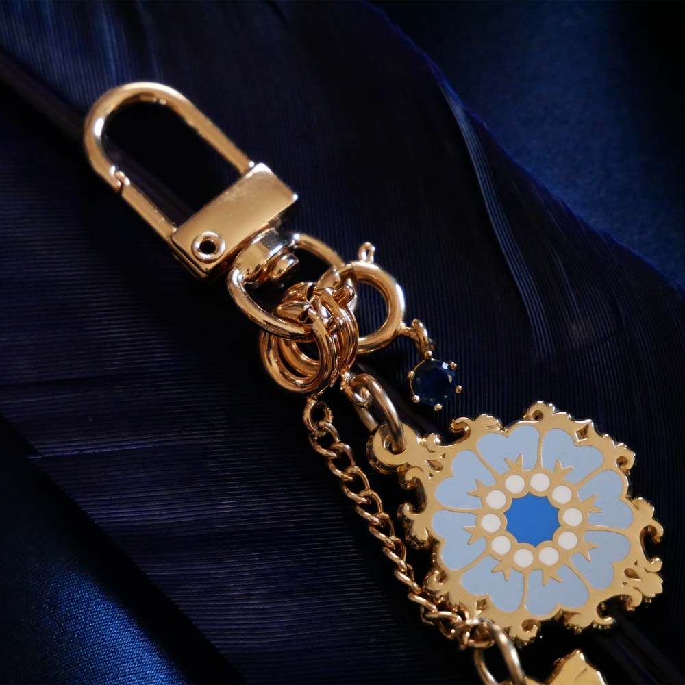 A detail shot of The Quirky Cup Collective Moncherie charmlet, an accessory with  a series of charms attached to a central clip. The design is gold and features a bow charm, a blue gem and a enamel element of a floral motif in a decorative french inspired style in shades of blue, cream and gold. The french inspired eReader accessory is placed on a royal blue fabric background on a matching coloured feather.