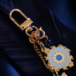 A detail shot of The Quirky Cup Collective Moncherie charmlet, an accessory with  a series of charms attached to a central clip. The design is gold and features a bow charm, a blue gem and a enamel element of a floral motif in a decorative french inspired style in shades of blue, cream and gold. The french inspired eReader accessory is placed on a royal blue fabric background on a matching coloured feather.