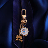 The Quirky Cup Collective Moncherie charmlet is is a series of charms attached to a central clip. The design is gold and features a bow charm, a blue gem and a enamel element of a floral motif in a decorative french inspired style in shades of blue, cream and gold. The french inspired eReader accessory is placed on a royal blue feather on a matching fabric background.