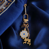 The Quirky Cup Collective Moncherie charmlet is is a series of charms attached to a central clip. The design is gold and features a bow charm, a blue gem and a enamel element of a floral motif in a decorative french inspired style in shades of blue, cream and gold. The french inspired eReader accessory is attached to a moncherie eReader case on a royal blue fabric background.