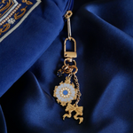 A detail shot of The Quirky Cup Collective Moncherie charmlet, an accessory with  a series of charms attached to a central clip. The design is gold and features a bow charm, a blue gem and a enamel element of a floral motif in a decorative french inspired style in shades of blue, cream and gold. The french inspired eReader accessory is attached to the zip of a matching moncherie eReader sleeve and sits on a matching royal blue background.