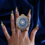 A hand with intricate gold and gem nails holds the moncherie clingie in front of a royal blue background. The kindle grip (similar to an enamel pop socket, also known as a clingie!) features a floral motif in a decorative french inspired style in shades of blue, cream and gold. The attachment device is in a light blue.