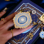 A hand with intricate gold and gem nails holds an ereader with a moncherie clingie (and matching moncherie charmlet and kindle insert sticker) in front of a royal blue background. The kindle grip (similar to an enamel pop socket, also known as a clingie!) features a floral motif in a decorative french inspired style in shades of blue, cream and gold. The attachment device is in a light blue.