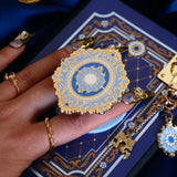 A hand with intricate gold and gem nails holds an ereader with a moncherie clingie (and matching moncherie charmlet and kindle insert sticker) in front of a royal blue background. The kindle grip (similar to an enamel pop socket, also known as a clingie!) features a floral motif in a decorative french inspired style in shades of blue, cream and gold. The attachment device is in a light blue.