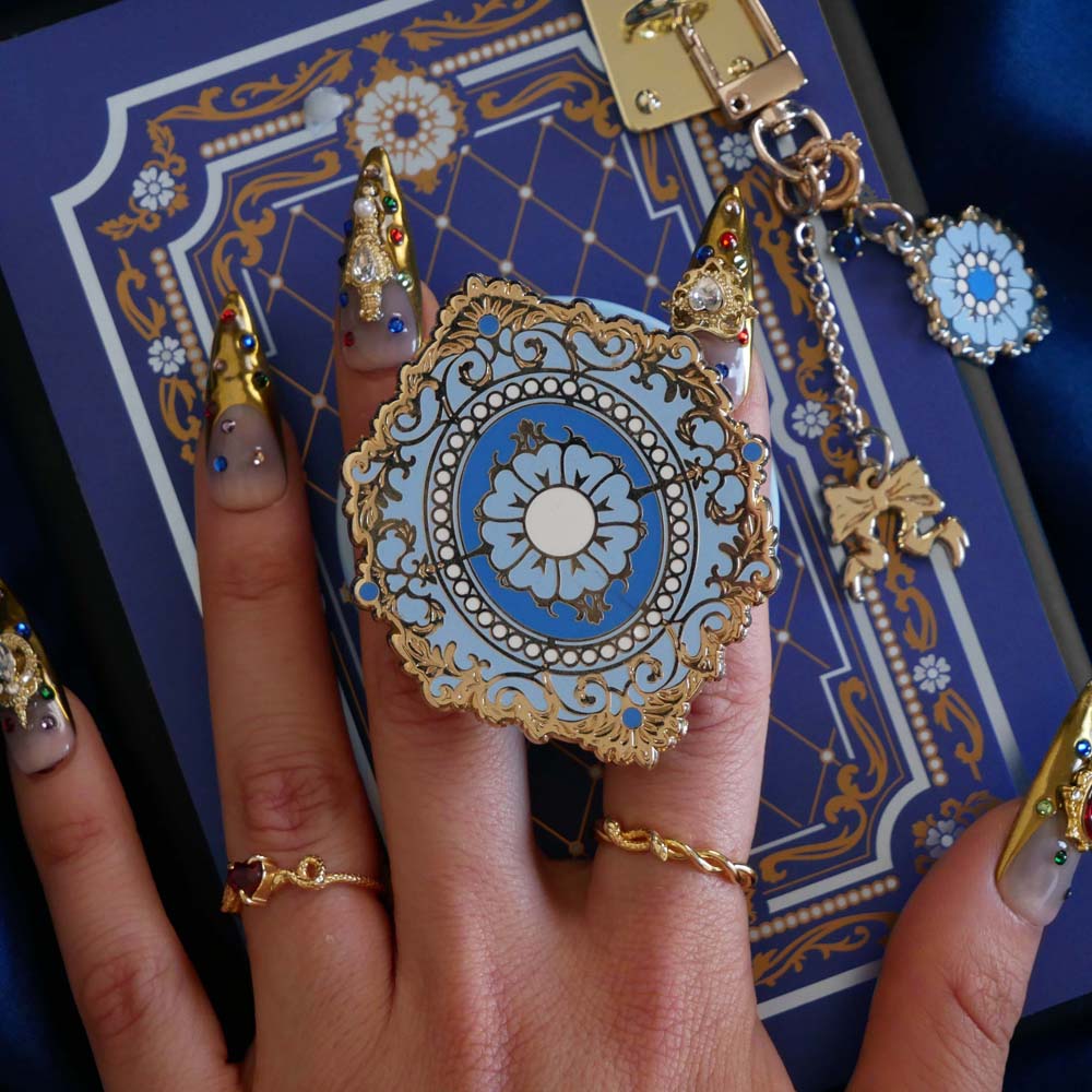 A hand with intricate gold and gem nails holds an ereader with a moncherie clingie (and matching moncherie charmlet and kindle insert sticker) in front of a royal blue background. The kindle grip (similar to an enamel pop socket, also known as a clingie!) features a floral motif in a decorative french inspired style in shades of blue, cream and gold. The attachment device is in a light blue.