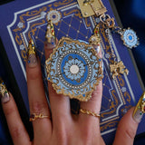 A hand with intricate gold and gem nails holds an ereader with a moncherie clingie (and matching moncherie charmlet and kindle insert sticker) in front of a royal blue background. The kindle grip (similar to an enamel pop socket, also known as a clingie!) features a floral motif in a decorative french inspired style in shades of blue, cream and gold. The attachment device is in a light blue.