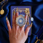 A hand with intricate gold and gem nails holds an ereader with a moncherie clingie (and matching moncherie charmlet and kindle insert sticker) in front of a royal blue background. The kindle grip (similar to an enamel pop socket, also known as a clingie!) features a floral motif in a decorative french inspired style in shades of blue, cream and gold. The attachment device is in a light blue.