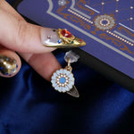 The Quirky Cup Collective Moncherie dust plug features a floral motif in a decorative french inspired style in shades of blue, cream and gold. The enamel element it attached to a transparent USB-c insert and is perfect for feminine kindle decor. The unique ereader accessory is in an ereader (with matching eReader insert sticker) and is held by a hand with intricate gold nails on a dark blue background.