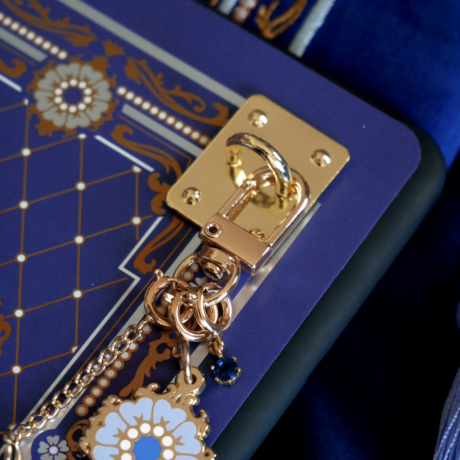 A detail shot of The Quirky Cup Collective Moncherie charmlet, an accessory with  a series of charms attached to a central clip. The design is gold and features a bow charm, a blue gem and a enamel element of a floral motif in a decorative french inspired style in shades of blue, cream and gold. The french inspired eReader accessory is attached to a kindle with a matching eReader sticker insert via a charm anchor, a plate with a clip for attaching accessories directly to your romantasy inspired ereader. 