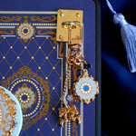 A detail shot of The Quirky Cup Collective Moncherie charmlet, an accessory with  a series of charms attached to a central clip. The design is gold and features a bow charm, a blue gem and a enamel element of a floral motif in a decorative french inspired style in shades of blue, cream and gold. The french inspired eReader accessory is attached to a kindle with a matching eReader sticker insert via a charm anchor, a plate with a clip for attaching accessories directly to your romantasy inspired ereader. 