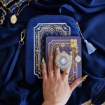 The Moncherie eReader decor bundle by The Quirky Cup Collective is the perfect feminine kindle aesthetic addition for book lovers. The set includes an eReader case, eReader sticker insert, charmlet, dust plug and clingie. The range features an intricate French inspired design in shades of gold, cream and light blue on dark blue backgrounds. The bundle is held by a hand with intricate gold nails on a royal blue fabric background, surrounded by gold bookish objects.