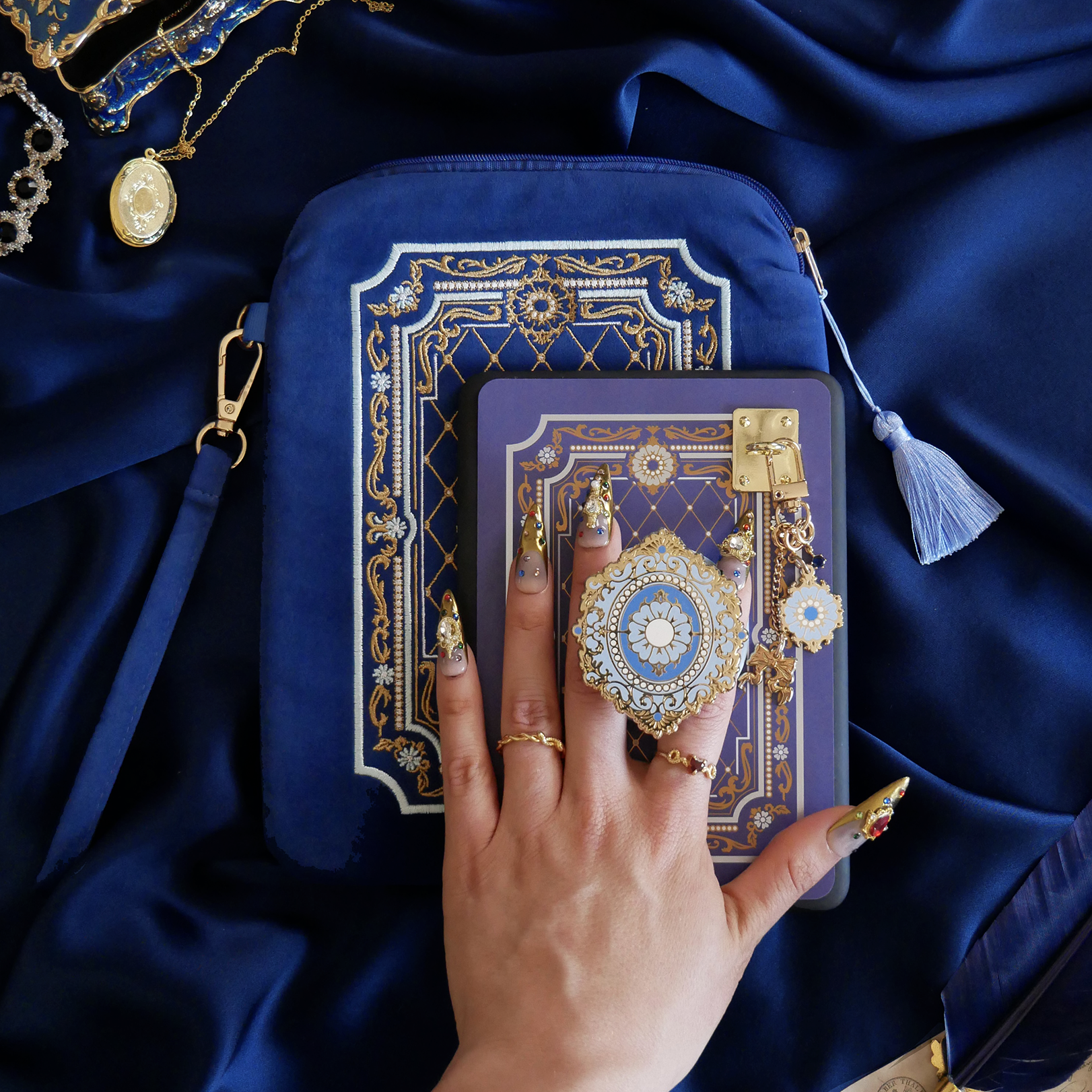 The Moncherie eReader decor bundle by The Quirky Cup Collective is the perfect feminine kindle aesthetic addition for book lovers. The set includes an eReader case, eReader sticker insert, charmlet, dust plug and clingie. The range features an intricate French inspired design in shades of gold, cream and light blue on dark blue backgrounds. The bundle is held by a hand with intricate gold nails on a royal blue fabric background, surrounded by gold bookish objects.