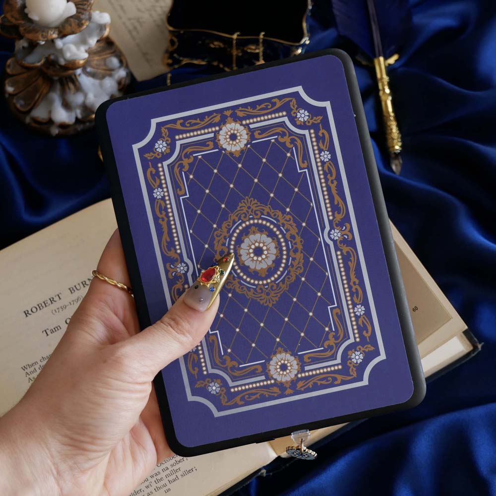 The moncherie eReader insert sticker is applied to an ereader and held in front of a classic book on a blue fabric background. The kindle sticker insert is a royal blue and features an intricate floral frame inspired design in shades of light blue, gold and cream.