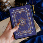 The moncherie eReader insert sticker is applied to an ereader and held in front of a classic book on a blue fabric background. The kindle sticker insert is a royal blue and features an intricate floral frame inspired design in shades of light blue, gold and cream.