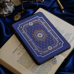 The moncherie eReader insert sticker is applied to an ereader that sits on a classic book on a blue fabric background. The kindle sticker insert is a royal blue and features an intricate floral frame inspired design in shades of light blue, gold and cream.