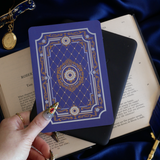 The moncherie eReader insert sticker is applied to an ereader and held in front of a classic book on a blue fabric background. The kindle sticker insert is a royal blue and features an intricate floral frame inspired design in shades of light blue, gold and cream.