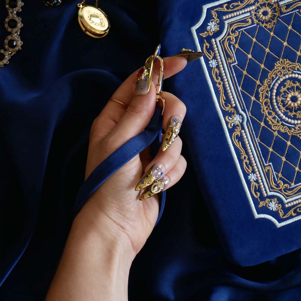 The Moncherie eReader and kindle case by The Quirky Cup Collective is the perfect feminine kindle aesthetic addition for book lovers. The blue velvet case features an intricate French inspired design in shades of gold, cream and light blue. The case also features a wrist strap with gold hardware, here being held by a hand with intricate gold nails. The case is styled on a royal blue fabric background and is surrounded by blue bookish props.