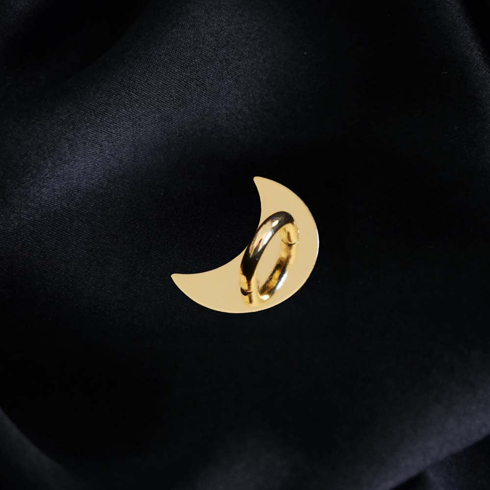 A The Quirky Cup Collective charm anchor on a black fabric background. The gold crescent moon-shaped clip is used to attach Charmlets to flat surfaces (such as your device). 