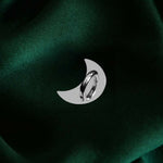 A The Quirky Cup Collective charm anchor on a green fabric background. The silver crescent moon-shaped clip is used to attach Charmlets to flat surfaces (such as your device).