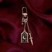 The  gold Night Realm Charmlet (A mix and match accessory for decorating your romantasy ereader aesthetic) by The Quirky Cup Collective sits on a dark red fabric background. The design featured a gold clasp and multiple charms hanging from different length detachable chains including an arched stained glass window with moon and star motifs, a sword and a small star.