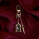 The  gold Night Realm Charmlet (A mix and match accessory for decorating your romantasy ereader aesthetic) by The Quirky Cup Collective sits on a dark red fabric background. The design featured a gold clasp and multiple charms hanging from different length detachable chains including an arched stained glass window with moon and star motifs, a sword and a small star.