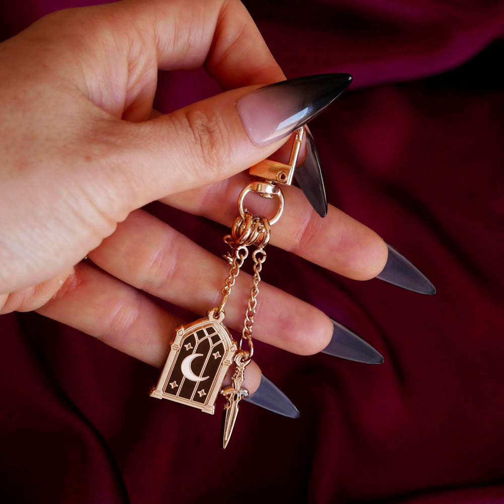 A detail shot of the gold Night Realm Charmlet (A mix and match accessory for decorating your romantasy kindle aesthetic) by The Quirky Cup Collective is held by a hand with long black nails over a dark red fabric background. The design featured a gold clasp and multiple charms hanging from different length detachable chains including an arched stained glass window with moon and star motifs, a sword and a small star.