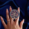 The blue oath Breaker clingie (think a bookish enamel pop socket!) is held in front of a royal blue background. The design centers on a crown, sword and snake motif with two hands at the bottom. The mostly circular (the bottom has a 'notch' Clingie is in shades of navy, teal, white and rich berry pink alongside the gold enamel.