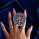 The blue oath Breaker clingie (think a bookish enamel pop socket!) is held in front of a royal blue background. The design centers on a crown, sword and snake motif with two hands at the bottom. The mostly circular (the bottom has a 'notch' Clingie is in shades of navy, teal, white and rich berry pink alongside the gold enamel.