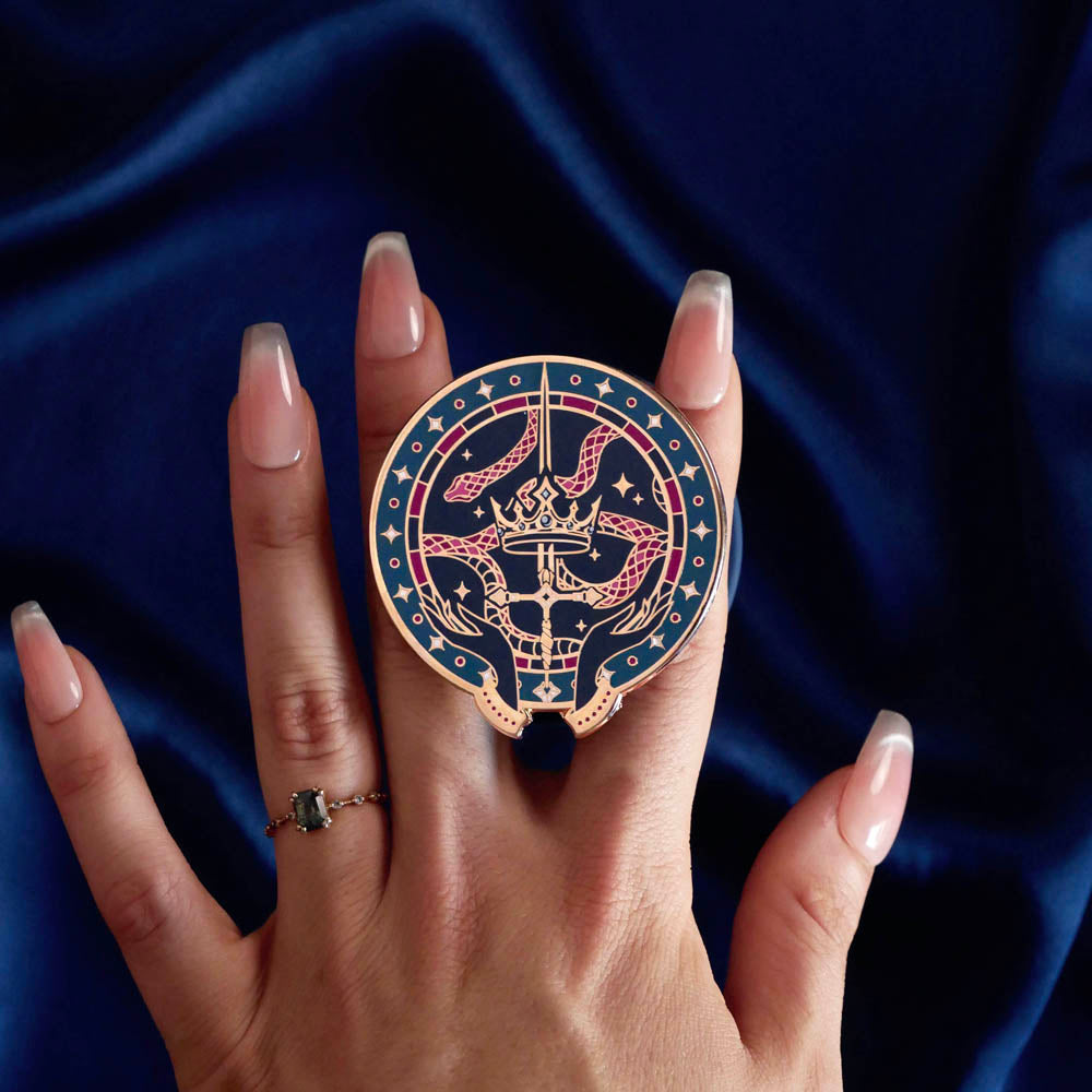 The blue oath Breaker clingie (think a bookish enamel pop socket!) is held in front of a royal blue background. The design centers on a crown, sword and snake motif with two hands at the bottom. The mostly circular (the bottom has a 'notch' Clingie is in shades of navy, teal, white and rich berry pink alongside the gold enamel.
