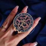 The blue oath Breaker clingie (think a bookish enamel pop socket!) is held in front of a royal blue background. The design centers on a crown, sword and snake motif with two hands at the bottom. The mostly circular (the bottom has a 'notch' Clingie is in shades of navy, teal, white and rich berry pink alongside the gold enamel.