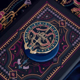 The blue oath Breaker clingie (think a bookish enamel pop socket!) is attached to a kindle with a matching navy eReader insert sticker. The Clingie design centers on a crown, sword and snake motif with two hands at the bottom. The mostly circular (the bottom has a 'notch' Clingie is in shades of navy, teal, white and rich berry pink alongside the gold enamel. The insert sticker features a similar design with more snakes and geometric detailing.