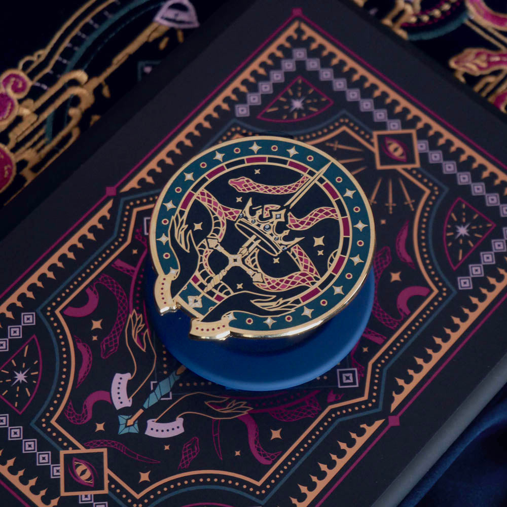 The blue oath Breaker clingie (think a bookish enamel pop socket!) is attached to a kindle with a matching navy eReader insert sticker. The Clingie design centers on a crown, sword and snake motif with two hands at the bottom. The mostly circular (the bottom has a 'notch' Clingie is in shades of navy, teal, white and rich berry pink alongside the gold enamel. The insert sticker features a similar design with more snakes and geometric detailing.