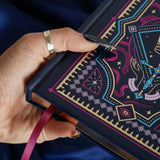 The navy blue Oath Breaker journal features an intricate cover foiled with shades of bright pink,, hold, teal and light pink. Motifs of snakes, crowns, swords, eyes and hands make for the perfect fantasy inspired gift. The journal sits on a royal blue fabric background to contrast the pink bookmark ribbon and gilded page edges.