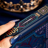 A detail of the spine of the navy blue Oath Breaker journal. the cover features intricate foiling in shades of bright pink,, hold, teal and light pink. Motifs of snakes, crowns, swords, eyes and hands make for the perfect fantasy inspired gift. The journal sits on a royal blue fabric background and is held by a hand.
