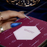 The internal nameplate of the blue Path Breaker journal sees a deep pink spread with snake and sword motifs. The journal is held by a hand on a dark blue fabric background. 