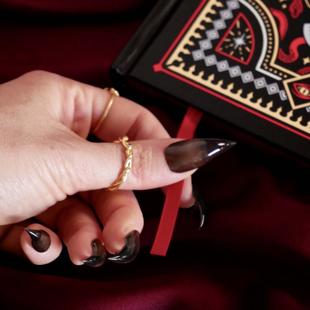 Hand with gold ring and black nail polish holding a red journal placeholder ribbon against a dark background