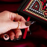 Hand with gold ring and black nail polish holding a red journal placeholder ribbon against a dark background