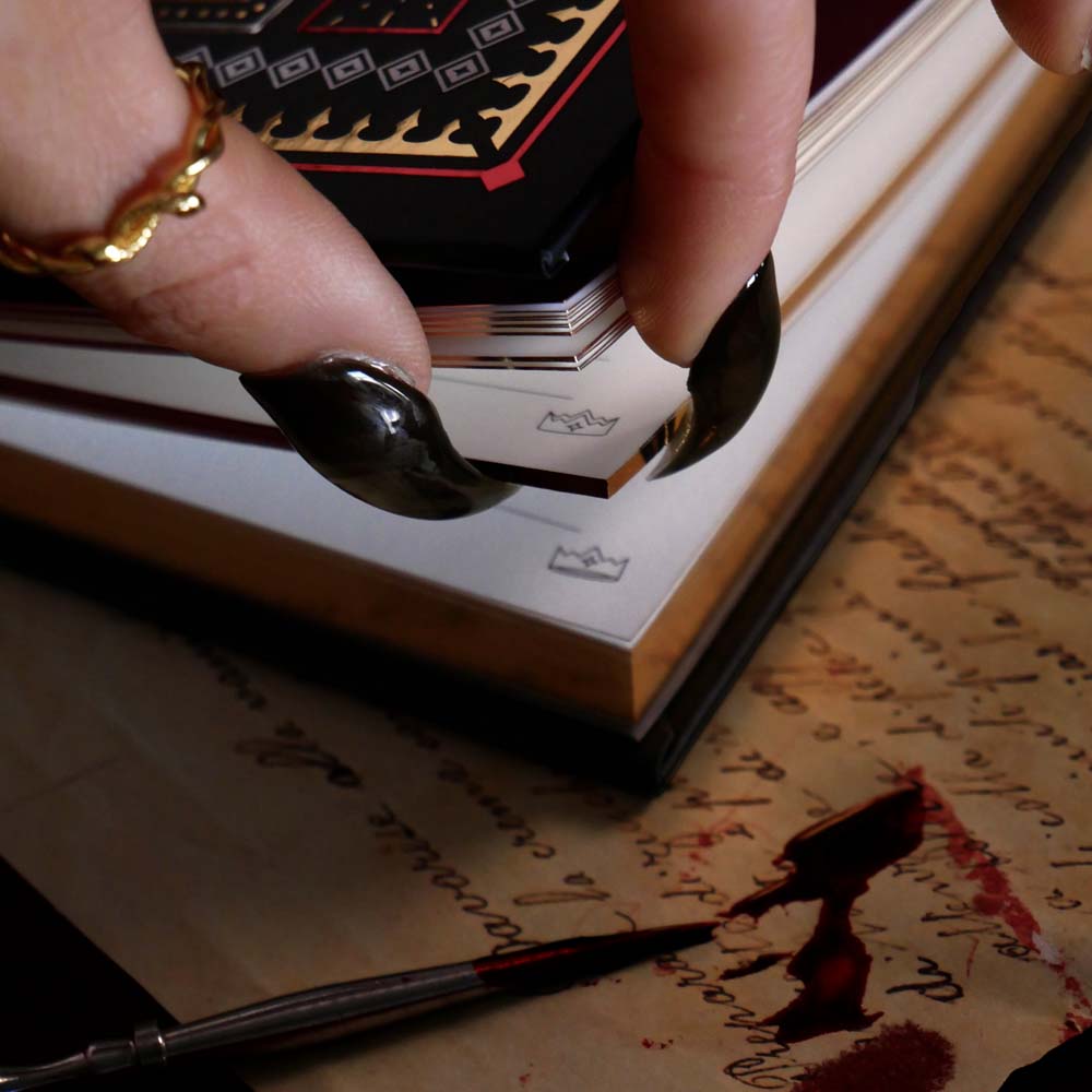 Close-up of a hand with black nail polish holding the corner of a page over an open notebook with writing on a wooden surface.