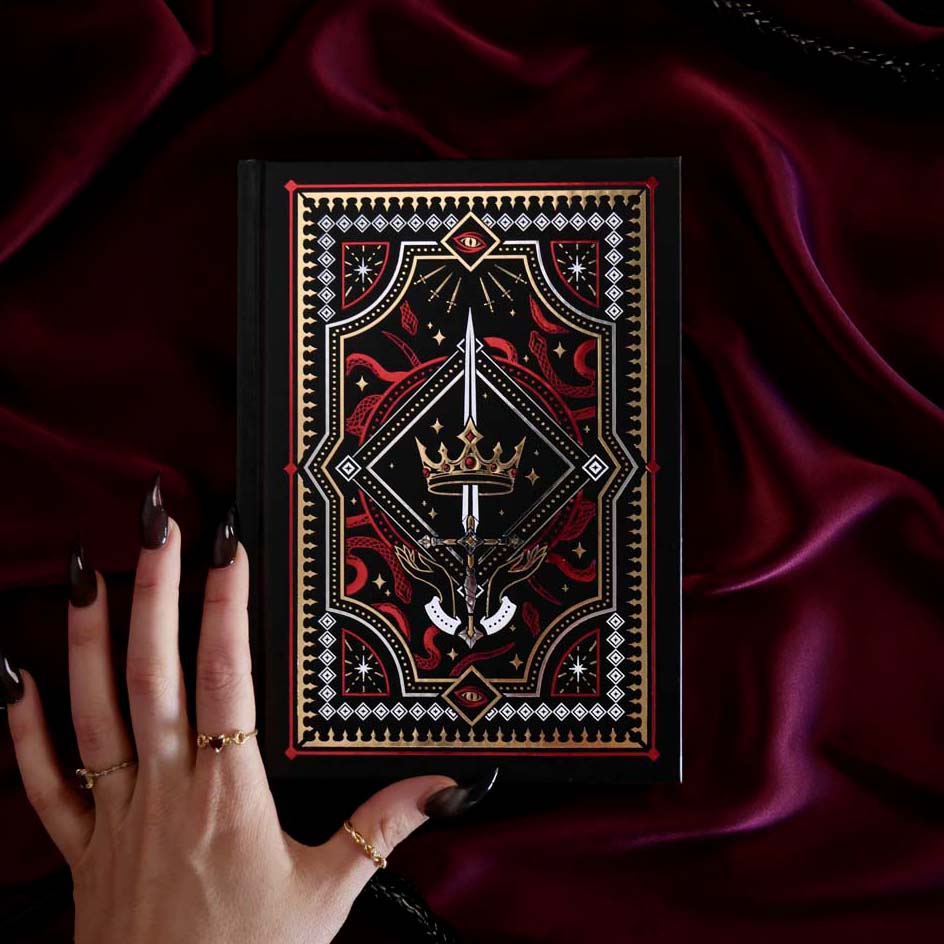 Decorative foiled journal with an intricate dark fantasy design held by a hand on a dark fabric background