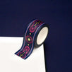 A close up of the Oath breaker washi tape by The Quirky Cup Collective. The roll of fantasy inspired washi tape is on the corner of a white journal on a blue background. The romantasy washi tape design is blue and pink with gold detailing- the intricate design showing a sword alongside geometric patterns.