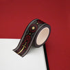 A close up of the Oath breaker washi tape by The Quirky Cup Collective. The roll of fantasy inspired washi tape is on the corner of a white journal on a red background. The romantasy washi tape design is red and black with gold detailing- the intricate design showing a sword alongside geometric patterns.