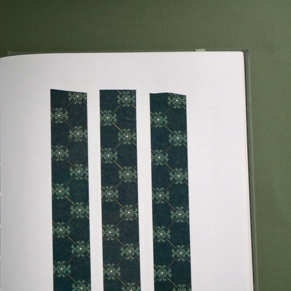 The Sage green variation of the Ornate Literati washi tape sits as three strips stuck to a white journal page on a green background with the ornate wallpaper inspired design in shades of dark green, sage green and gold.