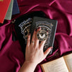 The black Otherworldly clingie by The Quirky Cup Collective is held in front of a matching kindle sticker insert and e-reader sleeve in front of a pink fabric background. The gold kindle grip features an intricate celestial inspired design of moons and stars in a minimal silver, gold, black and white colour palette.